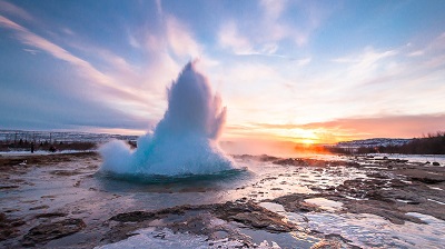 Geysern Strokkur skjuter upp en kaskad av kokhett vatten, mot en solnedgng med bl himmel och utdragna rosafrgade moln.